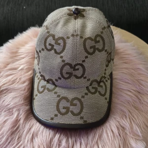 Gucci keps med GG-mönster - Snygg keps från Gucci i beige och brunt med det klassiska GG-logomönstret över hela kepsen. Skärmen har en mörkbrun kant i skinnimitation och kepsen är tillverkad i ett tåligt tyg. Perfekt accessoar för att lyfta din outfit.
