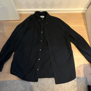 Svart skjorta från Weekday i bomull - Stilren svart skjorta från Weekday i storlek M. Skjortan har klassisk krage, knäppning framtill och en bröstficka. Tillverkad i mjuk bomull och har långa ärmar. Perfekt för dig som gillar enkel och clean stil. Funkar riktigt bra som en overshirt!