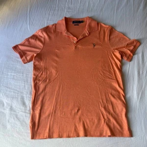 Orange pikétröja från Ralph Lauren - Snygg orange pikétröja från Polo Ralph Lauren i klassisk passform. Tröjan har korta ärmar, krage och knappslå framtill samt den ikoniska broderade loggan på bröstet. Perfekt för en chill och stilren look.