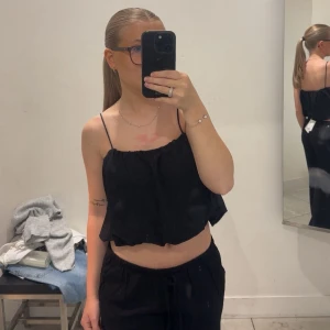 Svart croppad linnetopp med tunna band - Svart croppad linnetopp med tunna axelband och avslappnad passform. Toppen har volangig nederkant och är perfekt för varma dagar. Enkel och stilren design som passar till mycket. Helt ny och prislapp finns kvar
