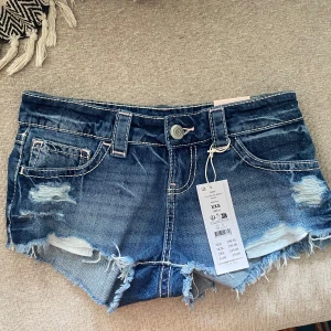 Lågmidjade jeansshorts från Gina Tricot - Snygga blå jeansshorts från Gina Tricot med låg midja, slitna detaljer och fransig kant. Shortsen har klassiska fickor fram och bak med coola sömmar och råa avslut. Perfekta för sommaren och ger en avslappnad vibe.