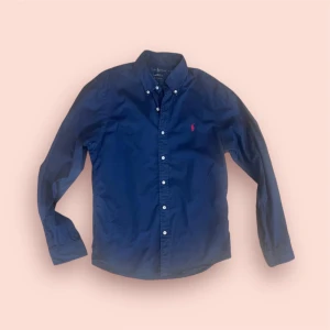 Mörkblå skjorta från Ralph Lauren - Klassisk mörkblå skjorta från Ralph Lauren med röd broderad logga på bröstet. Skjortan har slim fit, långa ärmar, knappar framtill och button-down krage. Tillverkad i bomull och passar perfekt för en clean och stilren look.