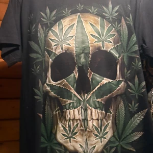 Svart t-shirt med dödskalle och cannabisblad - Cool svart t-shirt med stor dödskalle framtill, där hela motivet är gjort av gröna cannabisblad. T-shirten har korta ärmar och en klassisk passform. Perfekt för dig som gillar edgy och unika tryck. Alla tröjor som säljs är oanvända. Finns i strl S,L, XLL