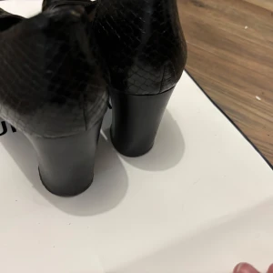 Svarta pumps med ormskinnsdetalj - Snygga svarta pumps med rund tå och hög klack. Skorna har en glansig finish framtill och coola ormskinnsdetaljer baktill som ger extra edge. Perfekta för dig som vill sticka ut med en klassisk men ändå trendig look.