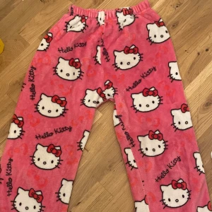 Hello Kitty rosa mjukisbyxor - Supergosiga rosa mjukisbyxor med Hello Kitty-motiv över hela byxorna. Byxorna har resår i midjan och är gjorda i mjuk fleece, perfekta för chill hemma. Sött tryck med Hello Kittys ansikte och röda rosetter.
