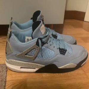Nike Air Jordan 4 University Blue - Nike Air Jordan 4 University Blue sneakers med ljusblå mocka, meshpaneler och grå detaljer med svarta stänk. Ikonisk Jordan -logga på hälen och tungans etikett. Skorna har snörning, svart och vit sula samt coola retrovibbar. Perfekta för dig som vill sticka ut Eller fån bra skott på basketplanen