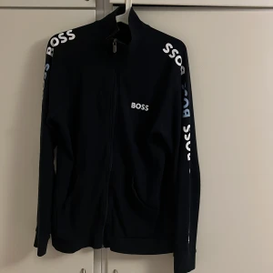 Mörkblå zip från BOSS - Mörkblå full zip från BOSS med logga tryckt på bröstet och längs båda ärmarna. Tröjan har hög krage, hel dragkedja och två fickor framtill. Perfekt för en sportig och clean look.