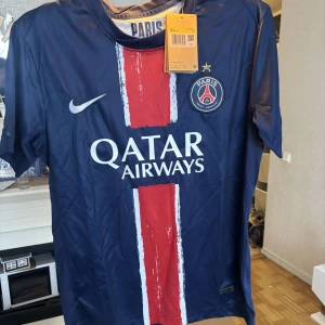 PSG Champion Edition 2024/25 - Paris Saint-Germain fotbollströja från Nike i marinblått med röd och vit rand längs mitten. PSG-emblem och Nike-logga på bröstet, samt Qatar Airways-tryck. Tillverkad i lätt och ventilerande material, perfekt för match eller träning.