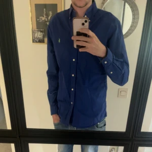 Blå skjorta från Ralph Lauren - Klassisk blå skjorta från Ralph Lauren med button-down krage och grönt broderat logotyp på bröstet. Skjortan har långa ärmar, knappar framtill och är tillverkad i bomull. Perfekt för en clean och stilren look.