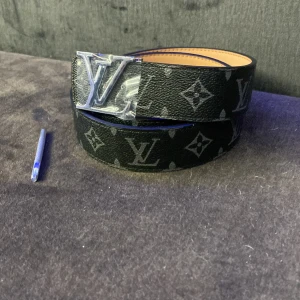 Louis Vuitton svart monogram bälte - Snyggt svart bälte från Louis Vuitton med klassiskt monogrammönster och stor LV-logga i silver på spännet. Bältet är gjort i skinn med beige insida och har exklusiv känsla. Perfekt accessoar för att lyfta din outfit.