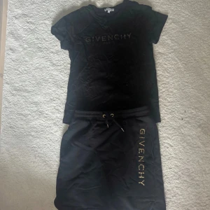 Svart Givenchy kjol med guldtext och matchande tröja - Supersnygg svart kortkjol från Givenchy med  resår i midjan och snörning. Kjolen har Givenchy-logga i guld längs ena sidan med coola spräckliga detaljer. Tröjan kommer med och matchar snyggt med likadan logga.  Perfekt för varje sommardag 