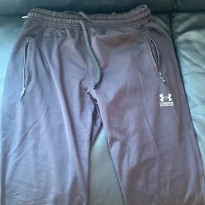 Svarta mjukisbyxor Under Armour L - Svarta mjukisbyxor från Under Armour med dragsko i midjan och dragkedjeförsedda fickor. Byxorna har logga på ena benet och är tillverkade i ett lätt och bekvämt syntetmaterial, perfekta för träning eller chill. Modellen är loose fit och har elastisk midja.