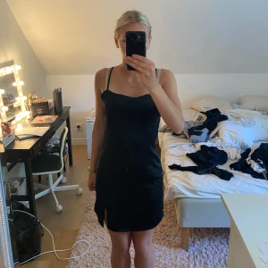 Svart satin klänning från BikBok - Supersnygg svart kort klänning från BikBok i satinliknande material. Den har smala axelband, en liten slits framtill och en enkel, stilren design som sitter figurnära. Perfekt för dig som gillar minimalistisk stil och vill ha något enkelt men ändå elegant. Dragkedja bak på den.