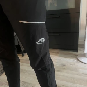 Svarta täckbyxor från The North Face - Säljer ett par svarta täckbyxor från The North Face med snygg logga på benet och en praktisk dragkedjeficka. Byxorna har en sportig look och är perfekta för kalla dagar. Materialet är slitstarkt och vattenavvisande, perfekt för utomhusaktiviteter.