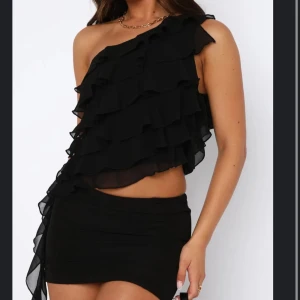 Svart one shoulder volangklänning - Supersnygg svart kortklänning med one shoulder och flera lager volanger upptill. Klänningen har en tight passform och är perfekt för dig som vill sticka ut. Tunnare, flowigt material som ger en cool look.