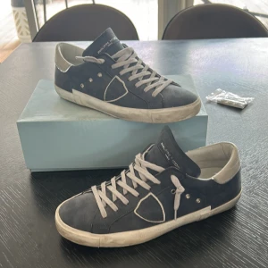 Philippe Model blå mockasneakers - Snygga sneakers från Philippe Model i mörkblå mocka med vita detaljer och grå snörning. Skorna har en klassisk låg profil, rund tå och vit sula med vintage-look. Ikonisk sköld på sidan och diskret branding på plösen. Perfekta för en stilren och avslappnad vibe.