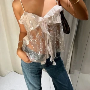 Glittrig volangtop med paljetter - Säljer en transparent glittrig topp med paljetter och volangkant. Toppen har tunna axelband, knytning framtill och är ärmlös. Perfekt för dig som vill sticka ut med en söt och trendig look.