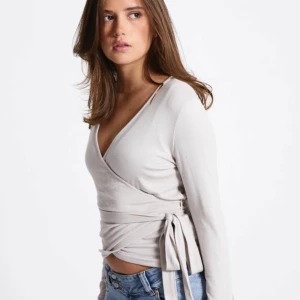 Beige omlottopp med knytband - Snygg beige omlottopp med långa ärmar och knytband i midjan. Toppen har v-ringad hals och sitter tajt runt kroppen för en stilren look. Perfekt att matcha med jeans eller kjol för en trendig outfit.