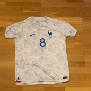 Frankrike Tchouaméni fotbollströja Nike - Snygg vit Frankrike fotbollströja från Nike med blå detaljer, nummer 8 och Tchouaméni på ryggen. Tröjan har korta ärmar och ett unikt ljusblått mönster med franska motiv. Perfekt för dig som vill sticka ut på match eller i vardagen.