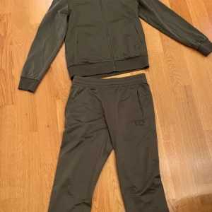 Olivgrön EA7 Emporio Armani tracksuit - Snygg olivgrön tracksuit från EA7 Emporio Armani. Setet består av en zip-jacka med hög krage och matchande byxor med resår i midjan. Båda delarna har diskreta fickor och EA7-logga tryckt på bröstet och benet. Perfekt för chill eller träning.