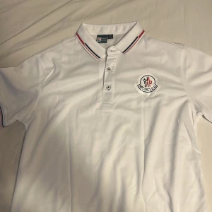 Vit Moncler pikétröja med logga - Snygg vit pikétröja från Moncler med klassisk krage och knäppning framtill. Tröjan har Moncler-logga på bröstet och diskreta rödblå ränder på krage och ärmslut. Perfekt för en clean och sportig look.