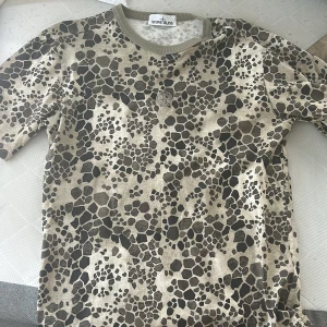 Limited edition alligator t-shirt Stone Island 2018 - Snygg t-shirt från Stone Island i beige med unikt alligatormönster i svart och grått. Klassisk rund halsringning med ribbad kant. Perfekt för dig som vill sticka ut med en streetig vibe och coolt mönster.                                                                    Kan köpas tillsammans med shortsen i ett set till ett bättre pris 