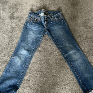 Blå jeans från American Eagle - Snygga blå jeans från American Eagle med klassisk femficksdesign och ljusa slitningar. Jeansen har raka ben och låg midja, samt coola detaljer på bakfickorna. Perfekta för en avslappnad och trendig look.