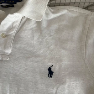 Vit pikétröja från Polo Ralph Lauren - Klassisk vit pikétröja från Polo Ralph Lauren i custom slim fit. Tröjan har korta ärmar, krage och knäppning framtill samt den ikoniska broderade loggan i mörkblått på bröstet. Perfekt för en clean och stilren look. Lite liten i storleken
