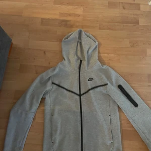 Grå Nike tech fleece - Snygg grå hoodie från Nike med hel dragkedja och svarta detaljer. Tröjan har huva, långa ärmar och en praktisk ficka på ena ärmen. Perfekt för en sportig look eller chill vardagsstil.
