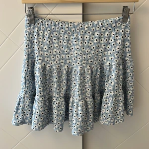 Blå blommig kortkjol - Säljer en söt blå kortkjol med vitt blommönster. Kjolen har en volangig nederdel och är lågmidjad. Knappt använd💕