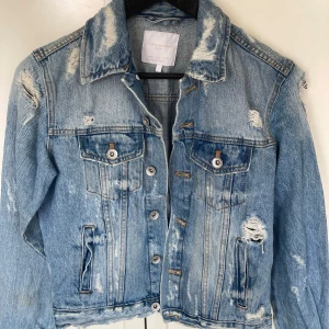 Ljusblå jeansjacka från Zara - Snygg ljusblå jeansjacka från Zara med slitna detaljer och klassisk krage. Jackan har knappar framtill, två bröstfickor med lock och två sidofickor. Perfekt för dig som gillar en avslappnad och trendig look.