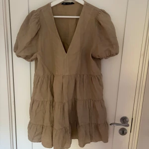 Beige kort klänning från Zara - Supersöt beige kortklänning från Zara med puffärmar och djup v-ringning. Klänningen har volanger i flera lager och är gjord i ett luftigt linnematerial, perfekt för varma dagar. Enkel men trendig design som ger en chill vibe.