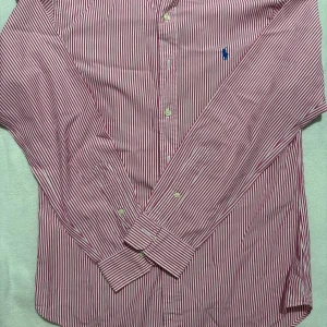 Randig skjorta från Polo Ralph Lauren - Snygg röd- och vitrandig skjorta från Polo Ralph Lauren i slim fit-modell. Skjortan har klassisk krage, långa ärmar och knäppning framtill. Broderad blå logga på bröstet ger en stilren touch. Perfekt för dig som gillar en fräsch och tidlös look.