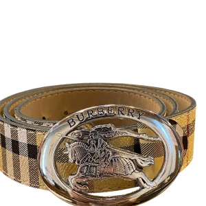 Rutig bälte med silverdetalj - Snyggt bälte med klassiskt rutigt mönster i beige, svart och brunt. Bältet har ett stort, ovalt spänne i silver  riddarmotiv. Tillverkat i skinn och perfekt för att lyfta vilken outfit som helst.