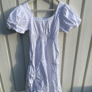 Vit långklänning med puffärmar - Superfin vit långklänning med puffiga kortärmar och smockad midja. Klänningen har en luftig och romantisk känsla med voluminösa ärmar och lätt rynkad kjol. Perfekt för sommarens alla tillfällen. Materialet känns svalt och skönt.