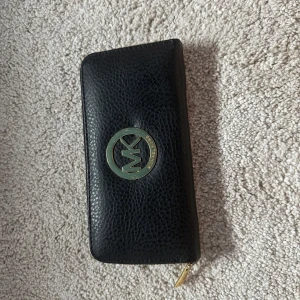 Svart plånbok från Michael Kors - Snygg svart plånbok från Michael Kors i skinn med guldigt MK-emblem framtill. Plånboken har dragkedja runtom, flera kortfack och innerfoder med MK-logga. Perfekt för att hålla koll på alla kort och cash med stil.