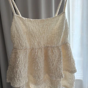 Vit volangtopp från Gina Tricot - Supersöt vit topp från Gina Tricot med smala axelband och volangig, rynkad struktur. Toppen har en luftig känsla och är perfekt för varma dagar. Materialet är lätt och känns som bomull eller syntet. Enkel och fräsch stil som passar till många tillfällen.