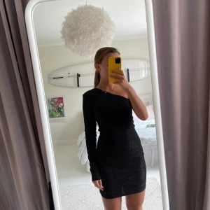 Svart glittrig one shoulder-klänning NA-KD - Säljer en svart glittrig kortklänning från NA-KD med one shoulder-design och lång ärm på ena sidan. Aldrig använd, som ny.