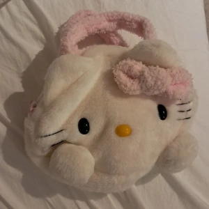 Hello Kitty fluffig handväska - Supergullig handväska officiell merch från Hello Kitty 2007 i fluffigt syntetmaterial. Väskan är formad som Hello Kittys ansikte i vitt med rosa detaljer och handtag, samt en söt rosett. Invändigt är den ljusrosa och stängs med dragkedja. Perfekt för dig som älskar sanrio