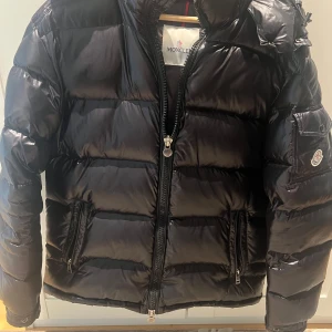 Svart Moncler pufferjacka med huva - Snygg svart pufferjacka från Moncler med glansig finish och klassisk logga på ärmen. Jackan har huva, två dragkedjefickor och kraftig dragkedja framtill. Perfekt för kalla dagar när du vill hålla stilen och värmen.