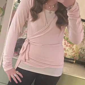 Ljusrosa omlottopp med knytband - Supersöt ljusrosa omlottopp med långa ärmar och knyt i sidan. Sitter snyggt figurnära och är perfekt att styla med jeans eller kjol. Mjuk och skön känsla, passar dig som gillar en feminin och enkel look.