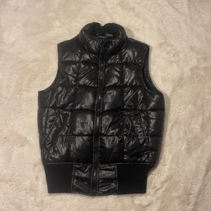 Svart dunväst från La Redoute - Snygg svart dunväst från La Redoute med glansig finish och quiltad design. Västen har hög krage, dragkedja framtill och två fickor med sned öppning. Perfekt att slänga över en hoodie för extra värme. Ribbad mudd nedtill för en schysst passform.