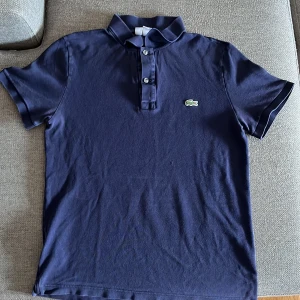 Mörkblå pikétröja Lacoste slim fit - Snygg mörkblå pikétröja från Lacoste i slim fit-modell. Klassisk krage, korta ärmar och knäppning med tre knappar framtill. Ikoniska gröna krokodilloggan broderad på bröstet. Tillverkad i mjuk bomull som känns skön mot huden. Original pris: 1100 kr fast jag säljer för 650 (kan gå ner till 600kr)