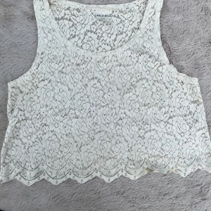 Vit spetsig linnetopp från Monki - Superfin vit linnetopp från Monki i spets med blommigt mönster. Croppad modell med ärmlös design och rund halsringning. Perfekt för sommarens alla tillfällen när du vill ha en romantisk och luftig vibe.