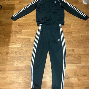 Adidas grönt träningsoverall med vita ränder - Säljer ett klassiskt grönt Adidas träningsset med vita ränder längs ärmar och ben. Setet består av en zip-jacka och matchande byxor, båda med Adidas logga på bröst och lår. Materialet är mjukt och stretchigt polyester, perfekt för sport eller chill.