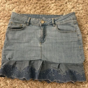 Jeanskjol med volang och spetskant - Snygg ljusblå jeanskjol från JAY med volang och broderad spetskant längst ner. Klassisk femficksmodell med knapp och dragkedja framtill. Perfekt för dig som gillar denim med en söt twist.