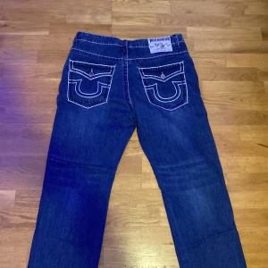 True Religion jeans blå - Säljer ett par klassiska True Religion Joey Super T jeans i storlek 38. Jeansen har kontrastsömmar, stora bakfickor med ikonisk broderad logga och raka ben. Tillverkade i USA och i 100% bomull. Oanvända.