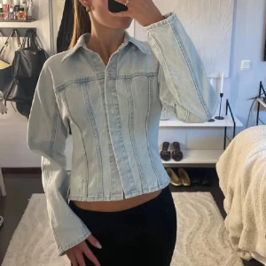 Ljusblå jeansjacka med vida ärmar - Säljer en snygg ljusblå jeansjacka med markerade sömmar och breda ärmar. Jackan har klassisk krage, knäppning framtill och en figurnära passform som ger en cool siluett. Perfekt att slänga över en t-shirt för en avslappnad look.