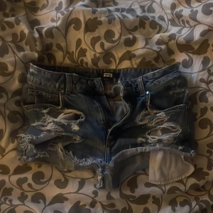 Rippade jeansshorts med nitar Bik Bok - Snygga blå jeansshorts från Bik Bok i storlek S. Shortsen har rippade detaljer framtill och coola guldfärgade nitar på ena bakfickan. Klassisk femficksmodell med låg midja och råa kanter. Perfekta för en edgy sommarlook!