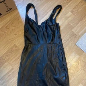 Svart klänning i skinnimitation NLY One - Snygg svart klänning från NLY One i skinnimitation med smala axelband och v-ringning. Klänningen är figurnära och har en kort längd, perfekt för dig som vill sticka ut. Enkel men edgy design som ger en cool vibe.